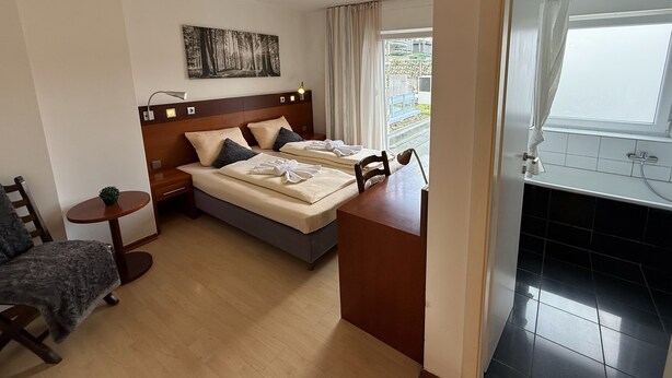 Une chambre d’hôtel avec un lit, des tables de chevet, une petite table ronde, une chaise, un tableau au mur et une fenêtre avec des rideaux.