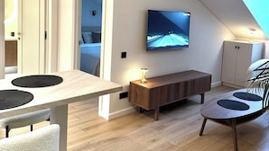 A modern living room with a wooden floor, a dining area with a table and chairs, a TV mounted on the wall, and a bench.