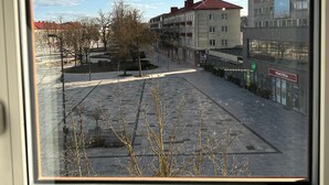 A view through a window of a building with a tiled plaza, leafless trees, and a clear sky.
