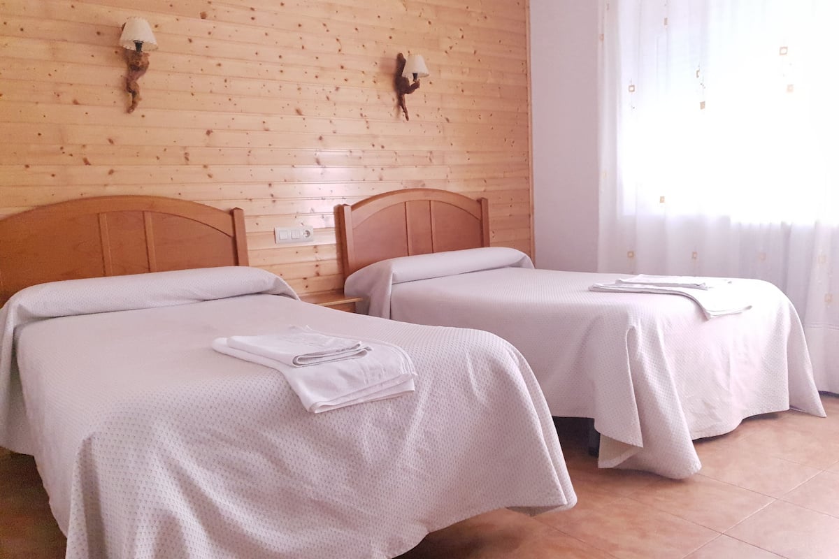 Two twin beds with white linens and towels, wooden headboards, and a wooden wall with two wall-mounted lamps.