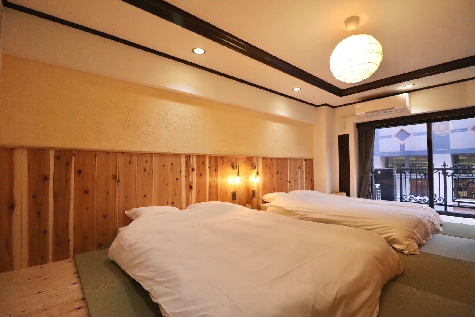 A hotel room with two beds, wooden paneling on the walls, a large window with a balcony, and a ceiling light fixture.