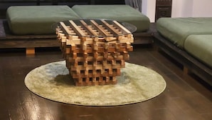 Una mesa redonda de madera con tapa de vidrio, rodeada por un tapete circular, en una habitación con suelo de madera y asientos tapizados en verde.