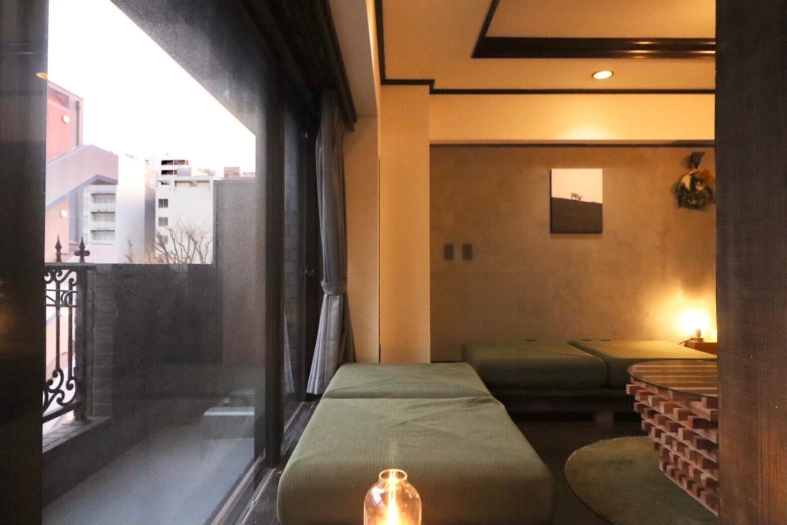 A room with a bed, a small table, a brick wall, and a window with a view of buildings.