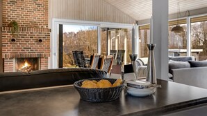 A modern living room with a brick fireplace, a large window, and a dining area with a basket of bread and plates.