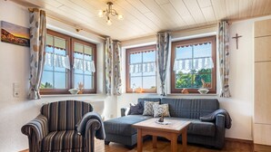 A cozy living room with a striped armchair, a sofa, a wooden coffee table, and large windows with curtains. - Apartment 'Sonnenblick, 54sqm' with Shared Terrace, Garden, and Wi-Fi (Schonach)