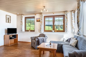 Une pièce avec un plafond en bois, un téléviseur posé sur un meuble en bois, un fauteuil à rayures, un canapé gris et une table sur laquelle se trouve un objet décoratif.