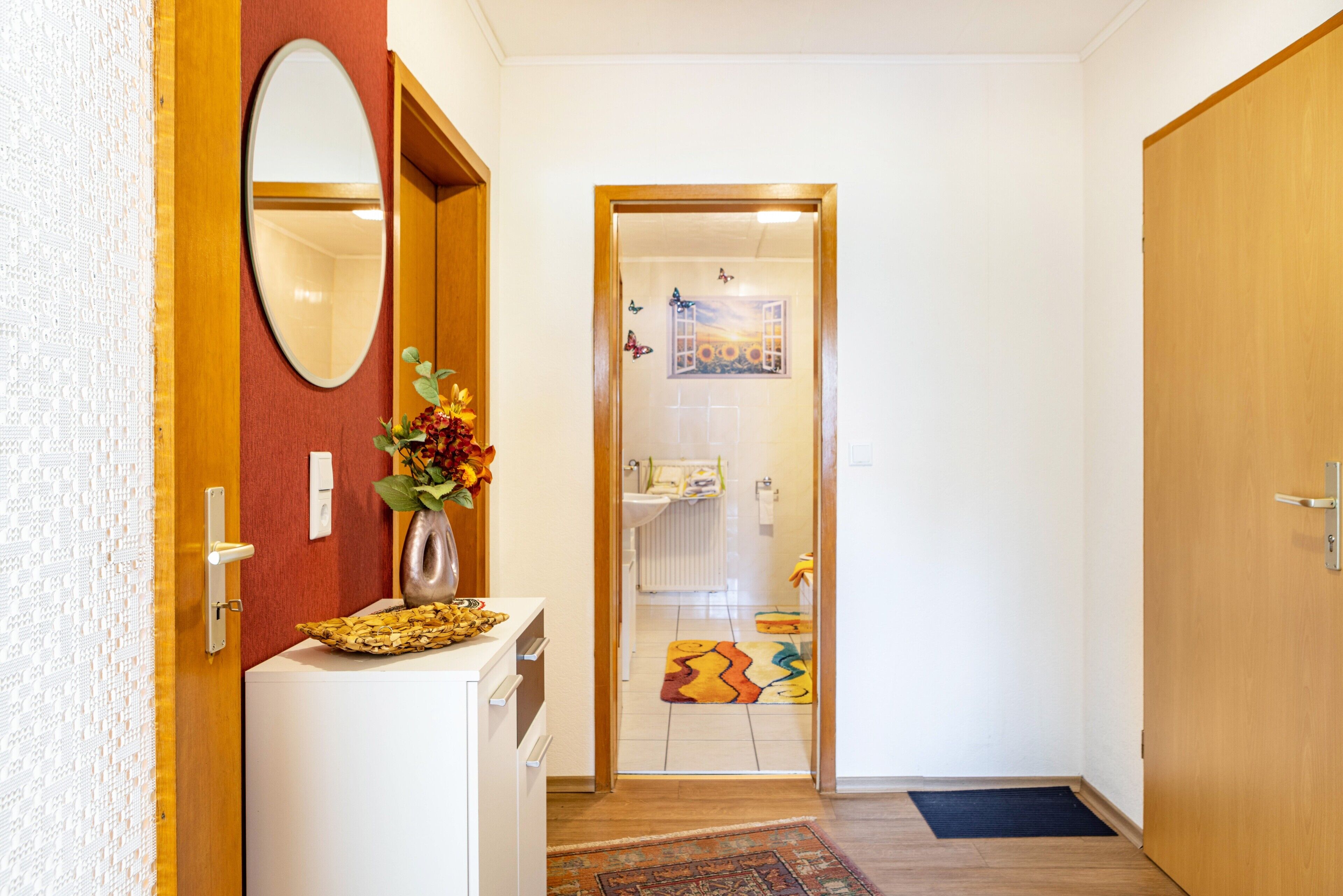 Un pasillo con una puerta de madera, un espejo, una mesita con un florero y una alfombra colorida que conduce a un baño.