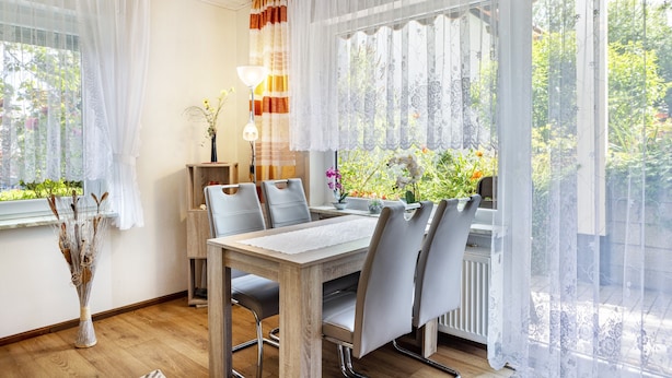 A dining area with a table, chairs, a vase with dried plants, and a window with curtains.