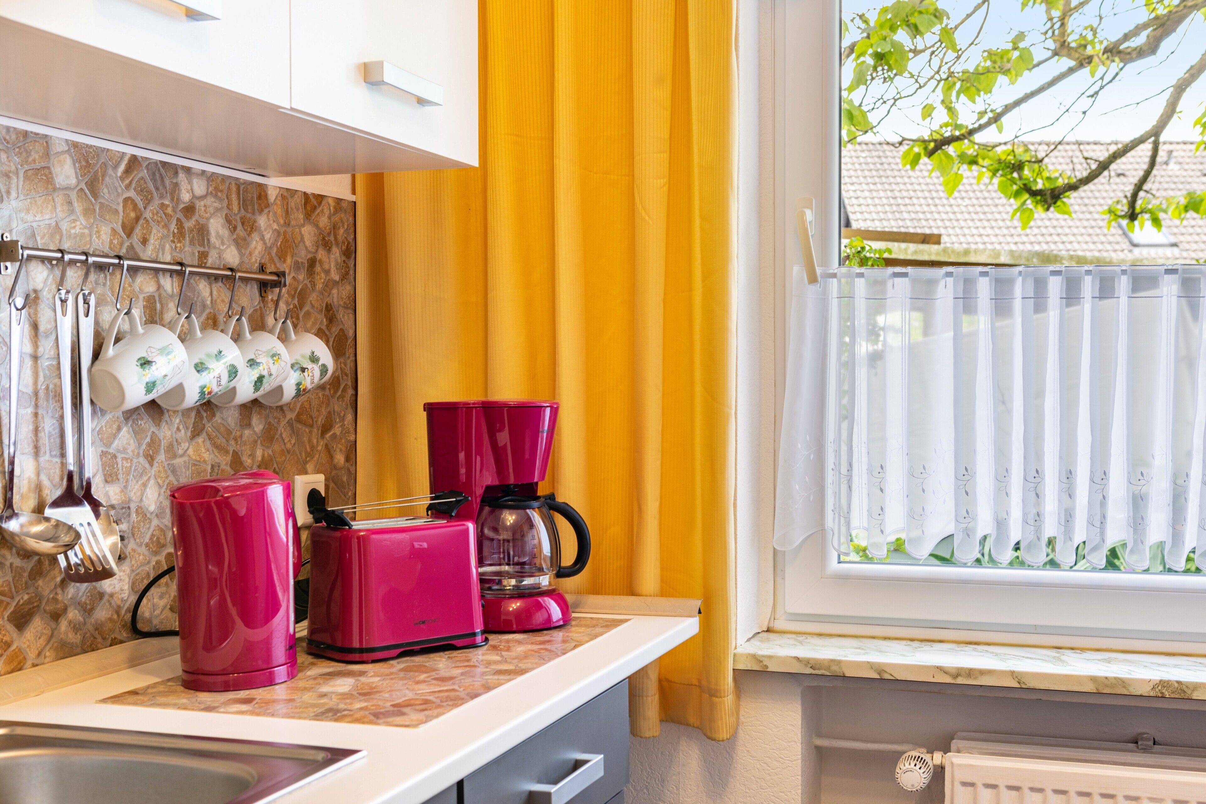 Eine Küche mit Steinrückwand, einem Fenster mit Blick auf einen Baum und einer Arbeitsplatte mit Kaffeemaschine und Toaster.