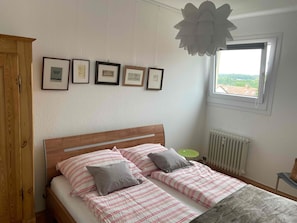 A bedroom with a wooden headboard, a bed with striped bedding, a radiator, a window with a view, and framed pictures on the wall.