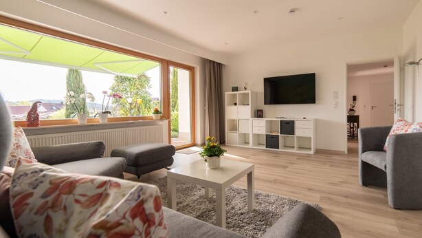 A modern living room with a sofa, a coffee table, a TV, and a large window with a view of greenery. - Apartment 'Pusteblume' with Private Terrace and Wi-Fi (Kappelrodeck)