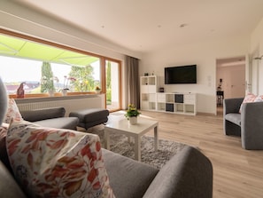 Een moderne woonkamer met een bank, een salontafel, een televisie en een groot raam met uitzicht op groen.