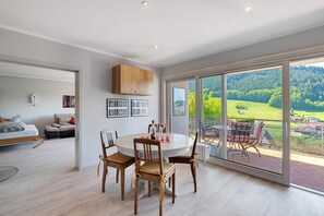 Een modern interieur met een eethoek, een bank en uitzicht op een groen landschap door grote ramen.