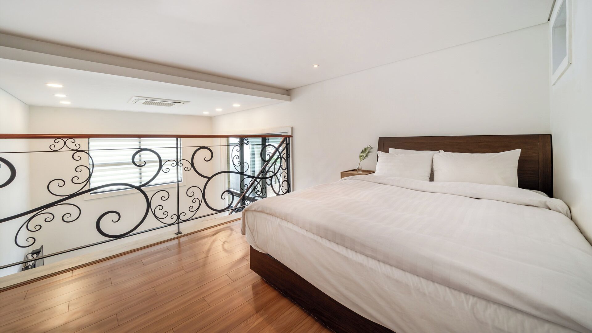 A bedroom with a large bed, wooden flooring, a balcony with decorative railing, and a ceiling light.