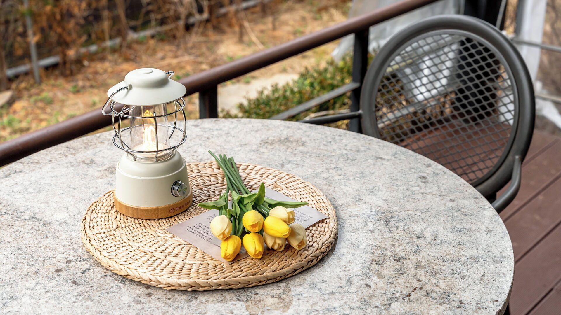 A round table with a woven placemat, a lantern, and a bouquet of yellow flowers.