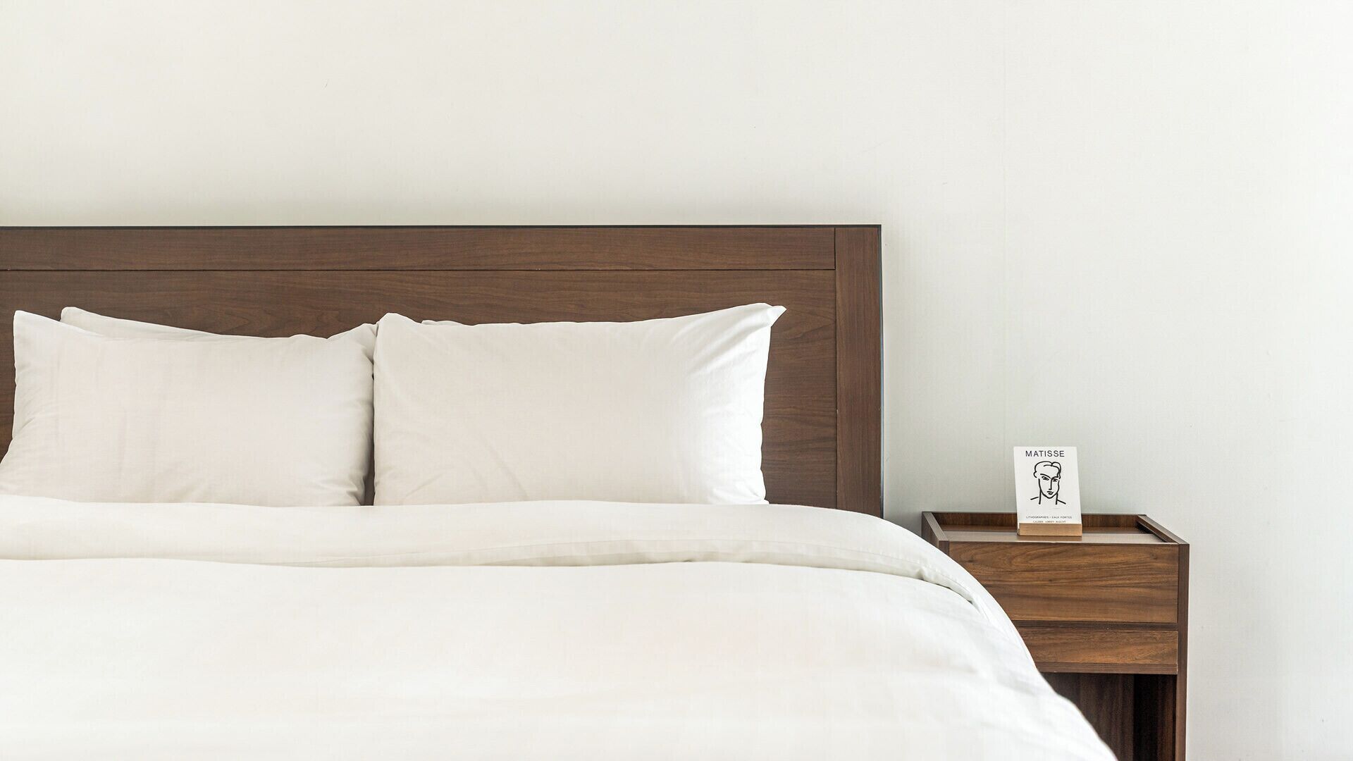 A bed with white bedding and two pillows, a wooden headboard, and a nightstand with a book titled 'Matisse' beside it.
