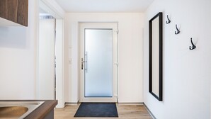 Un pasillo con una puerta de vidrio esmerilado, un felpudo negro en la entrada y un espejo con ganchos en la pared.
