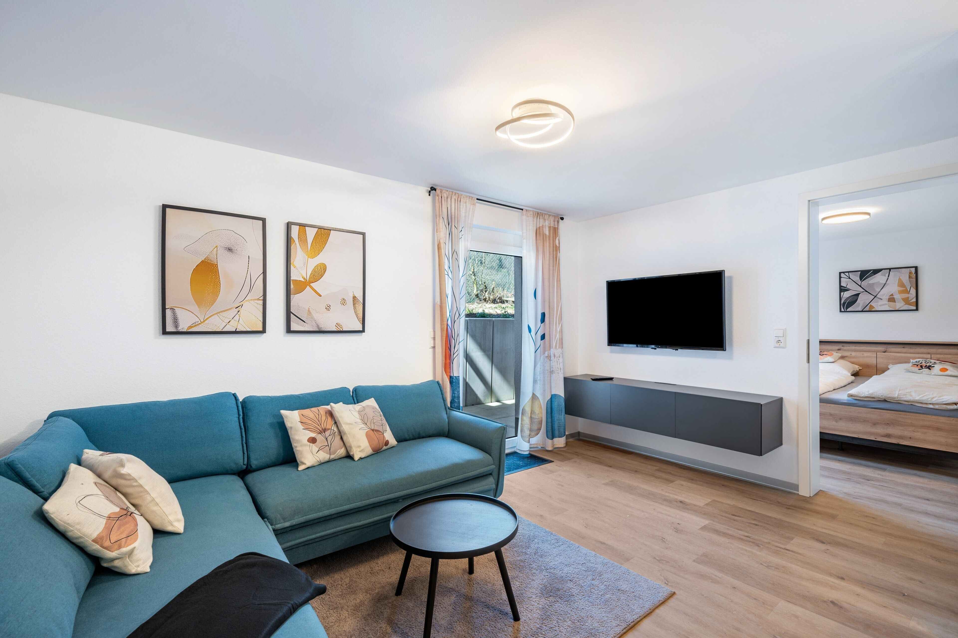 A modern living room with a sofa, a small round table, a TV mounted on the wall, and a bed with a headboard.