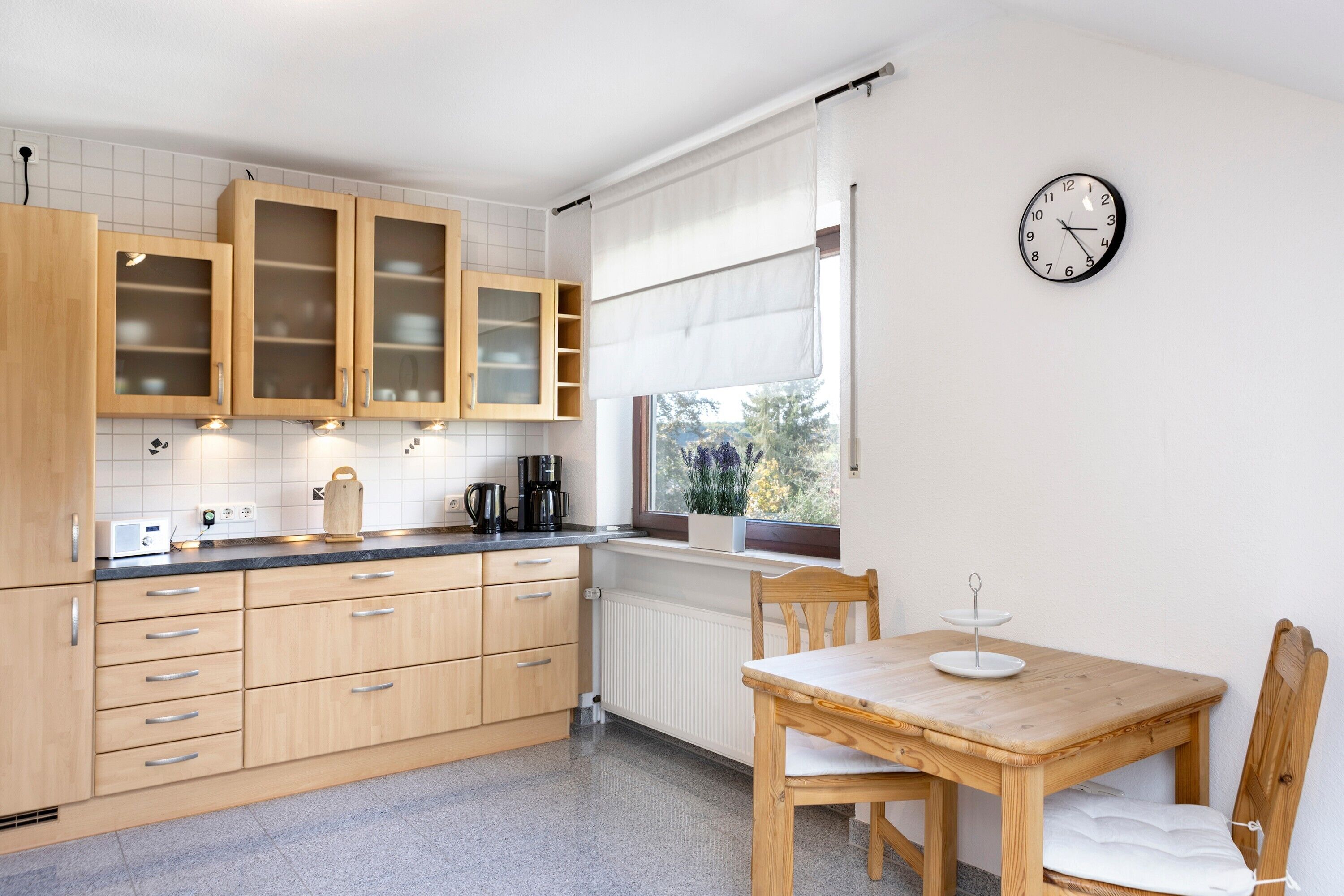 Eine Küche mit Holzausstattung, einem Fenster mit Vorhängen, einer Uhr und einem Esstisch mit Stühlen.