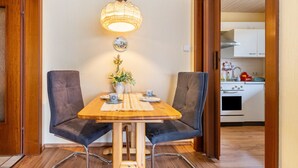 Un'area pranzo con un tavolo in legno, due sedie, un lampadario, un vaso con fiori e una cucina sullo sfondo.