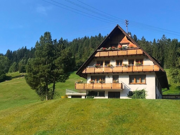 A two-story chalet with a sloped roof, wooden balconies, and a view of a forested hillside.