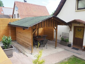 Et udendørs opholdsområde med træbænk, bord og stole, en lille overdækket konstruktion og en dør med en krans.
