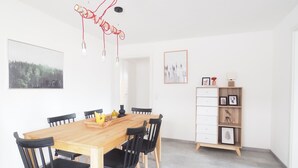 Et spiseområde med et træbord og sorte stole, en hvid væg med indrammede billeder og et træskab med skuffer.
