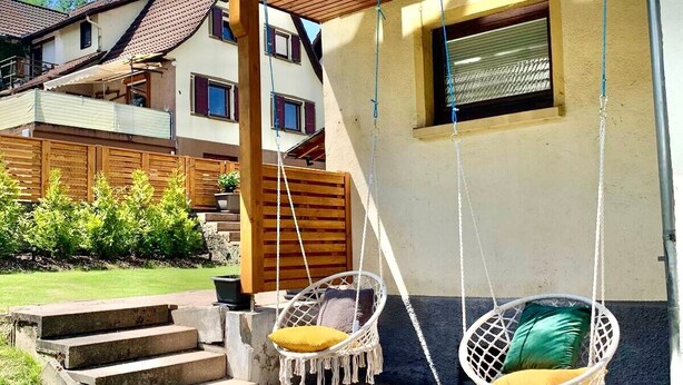 En boligbygning med en patio, to hængekøjsstole med hynder, en trappe af beton til indgangen og et træhegn.