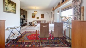 Een kamer met een eettafel, twee stoelen, een kamerkleed met patroon, een boekenkast en een groot raam.