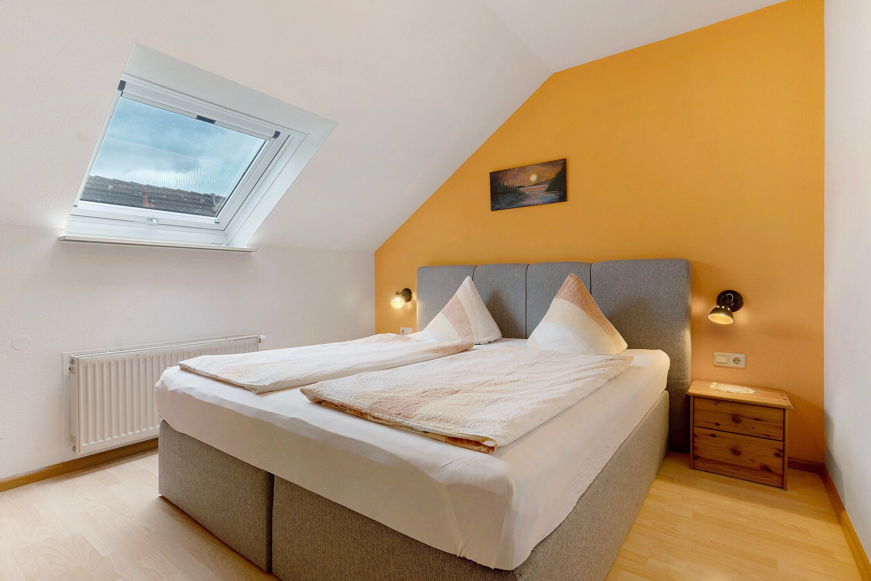 Quarto com claraboia, cama com cabeceira cinza, luminárias de cabeceira, mesa de cabeceira de madeira e radiador.