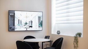 A modern hotel room with a round table and four black chairs, a large framed picture on the wall, and a window with blinds.