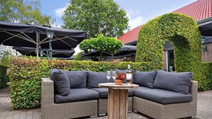 Een terras met twee grijze wickerbanken, een ronde houten tafel met een vaas en wijnglazen, en een bakstenen gebouw met een rood dak en beplanting.