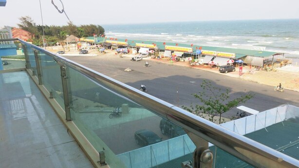 A coastal view from a balcony with a glass railing overlooking a beach and a road with vehicles and market stalls. - Minh Hanh Hotel Sam Son -BAY LUXURY (Sam Son)