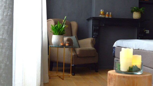 Una habitación con un sillón color beige, una pequeña mesa con una planta, una vela y un taburete de madera.