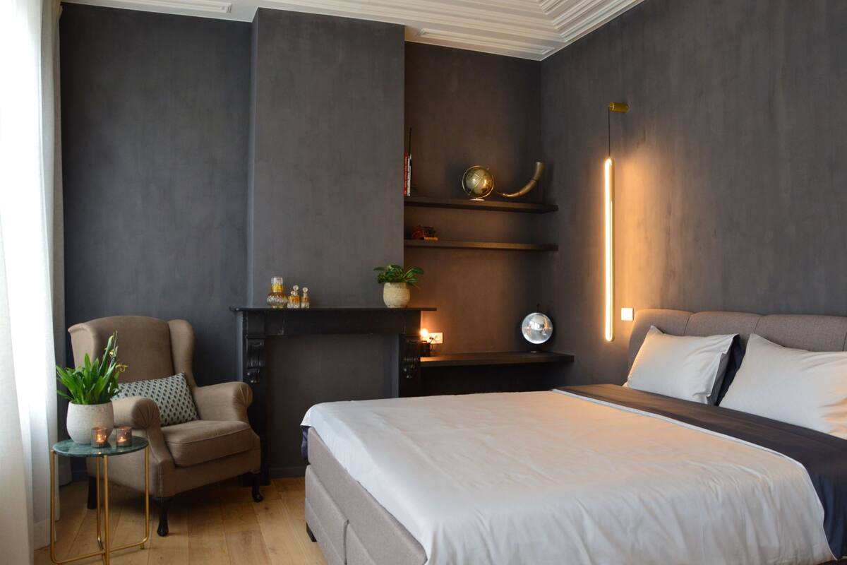 Un dormitorio moderno con una cama grande, una silla, una mesita con una planta y una estantería con adornos.