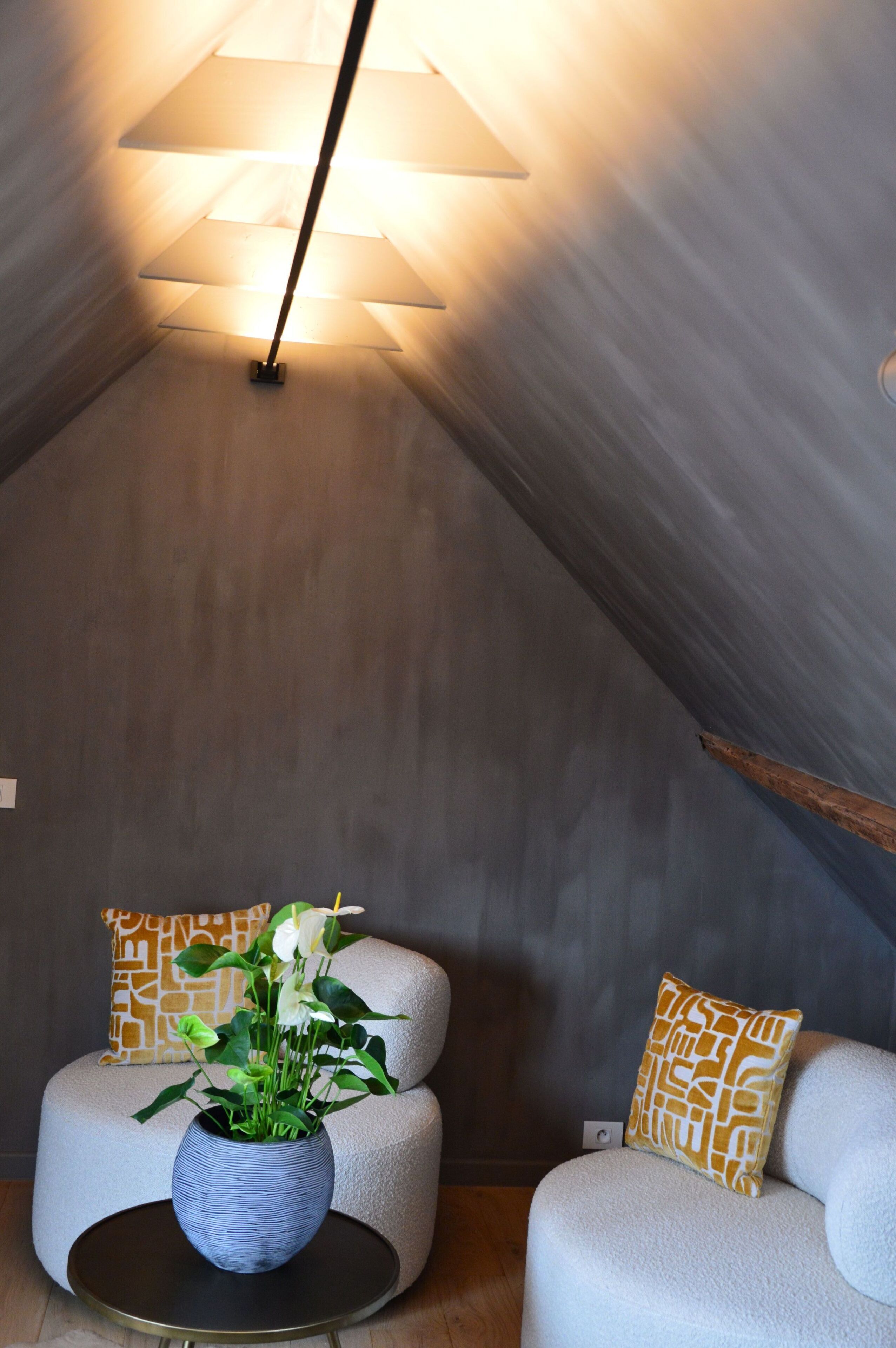A cozy room with a sloped ceiling, a round table with a vase of flowers, and two cushioned chairs with patterned pillows.