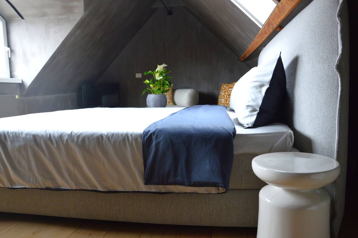 Un dormitorio con techo inclinado, una cama con manta azul, una mesita de noche blanca y una planta en maceta.