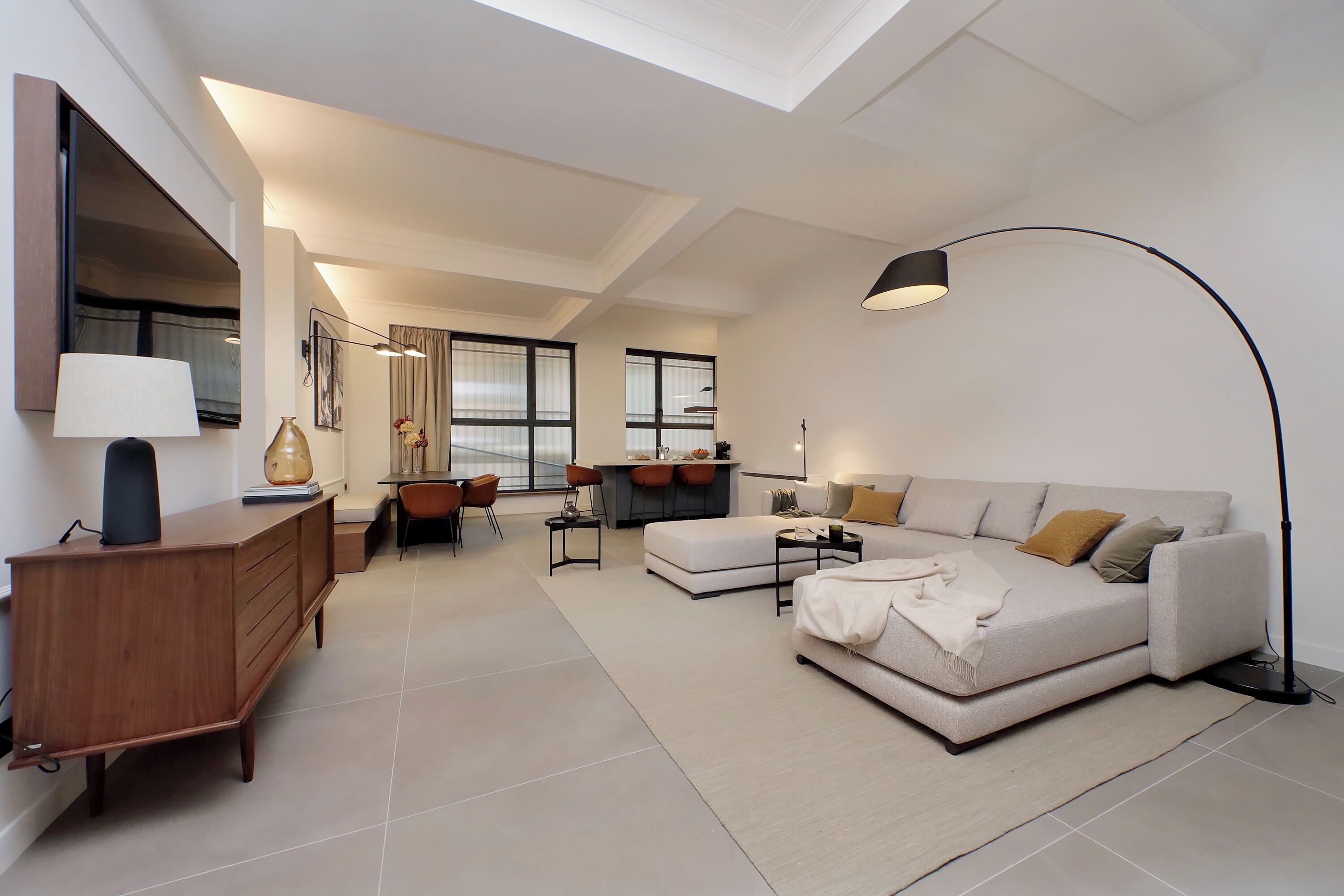 A modern living room with a sofa, a coffee table, a desk, and a chair. There is a large window providing natural light and a view of the outdoors.