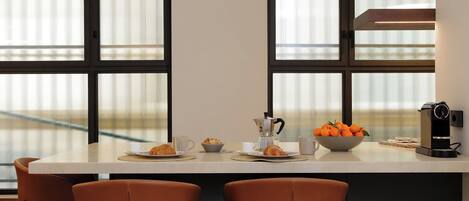 A modern kitchen with a central island, two brown chairs, a coffee maker, a bowl of oranges, and a window with blinds.