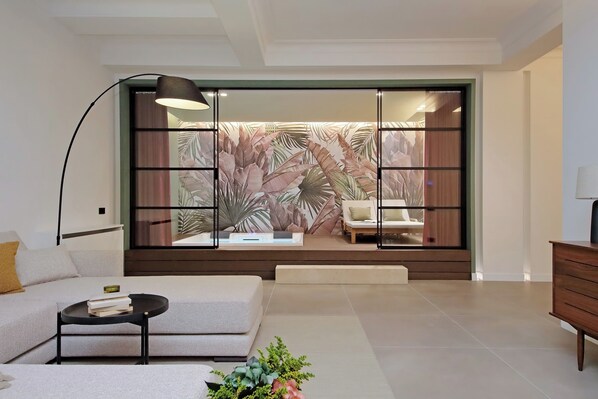 A modern living room with a sofa, a coffee table, a lamp, and a sliding glass door with a tropical leaf wallpaper. - Crispi Loft (Rome)
