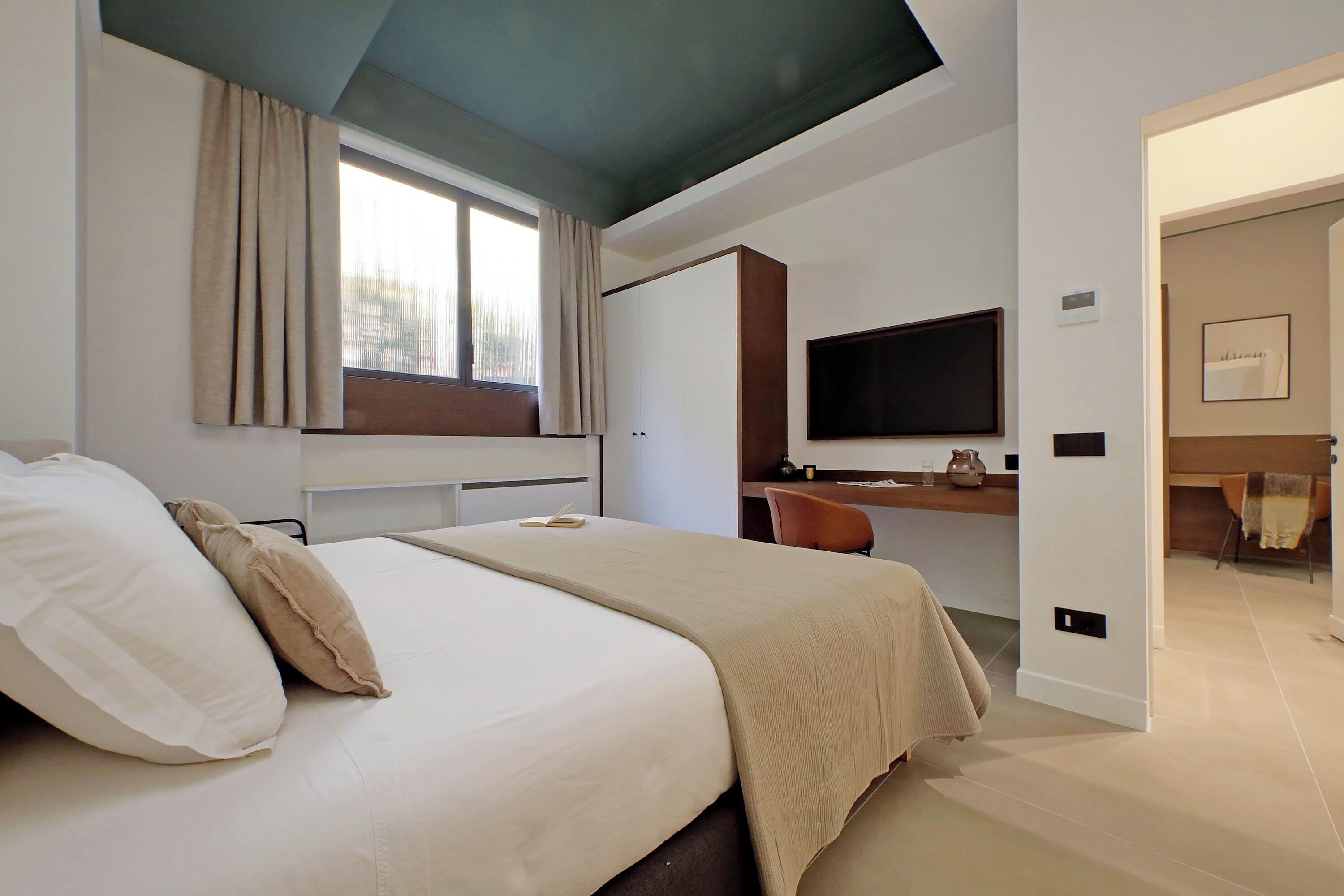 A modern hotel room with a large bed, a desk, a flat-screen TV, and a view of the outdoors.