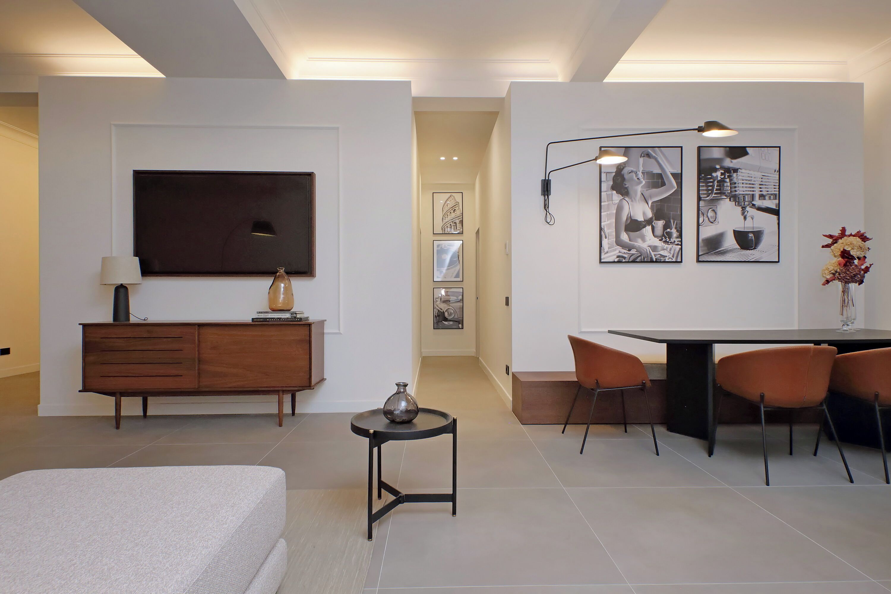 Quarto de hotel moderno com uma cama, uma pequena mesa com um vaso, uma TV, área de refeições com cadeiras e obras de arte nas paredes.