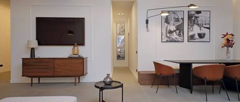 Quarto de hotel moderno com uma cama, uma pequena mesa com um vaso, uma TV, área de refeições com cadeiras e obras de arte nas paredes.