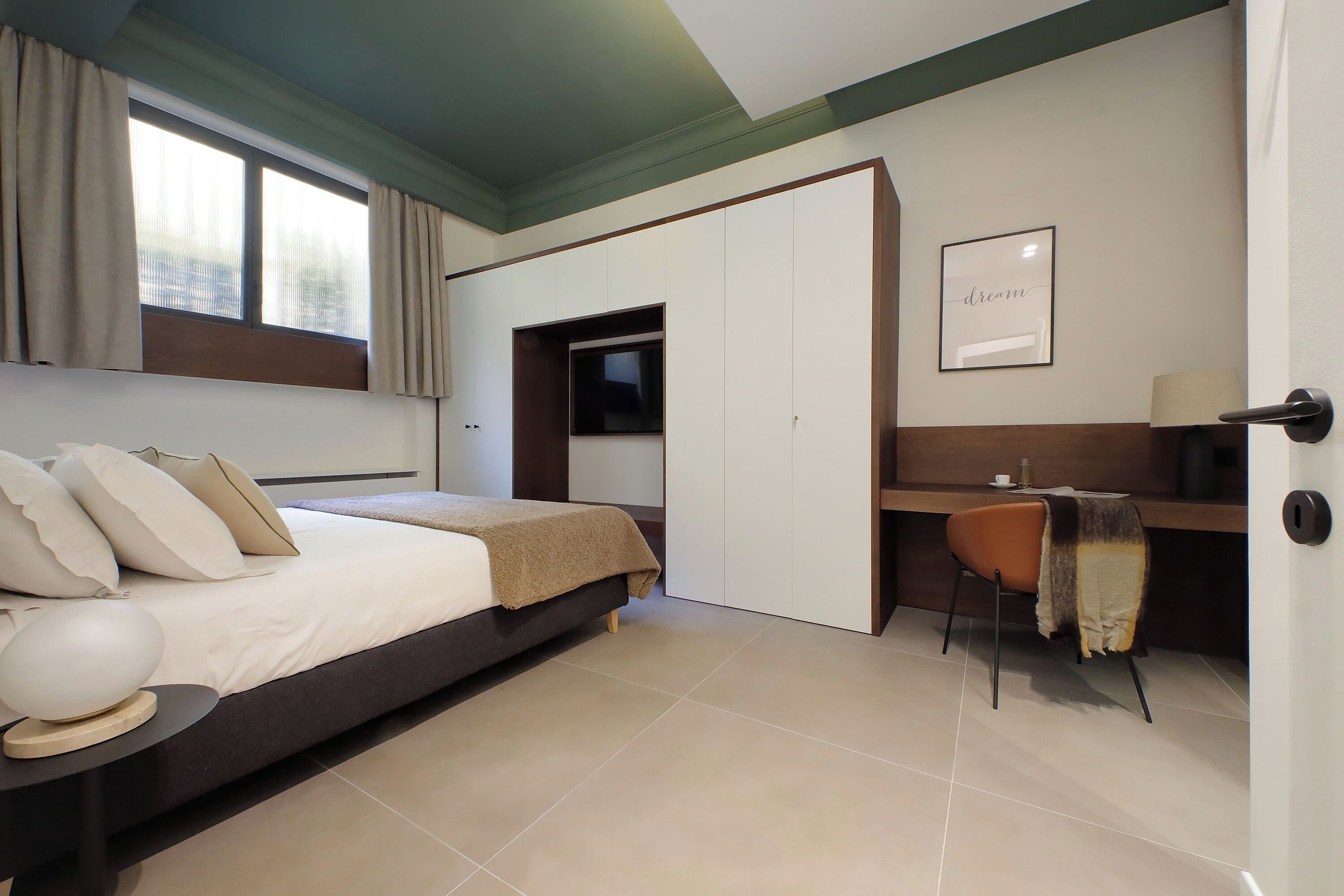 A modern bedroom with a bed, a desk, and a chair. There is a window with curtains, a framed picture on the wall, and a door.