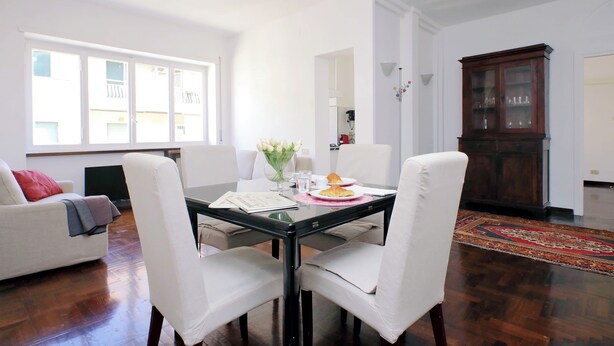 Un coin repas comprenant une table à plateau de verre, des chaises à revêtement blanc, un meuble en bois et une vue sur l’extérieur grâce à de grandes fenêtres.