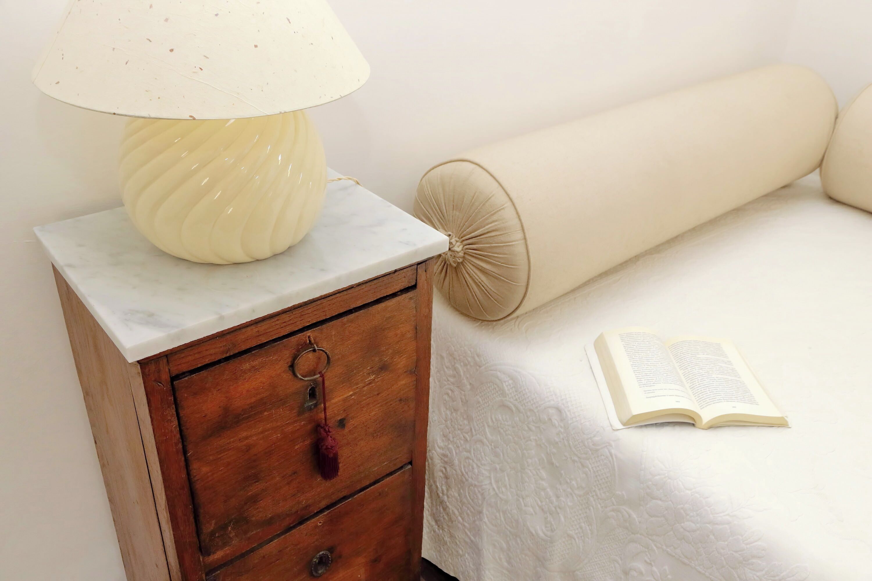 A bedside table with a lamp, a wooden nightstand drawer, a rolled-up beige blanket, and an open book on a white bed.