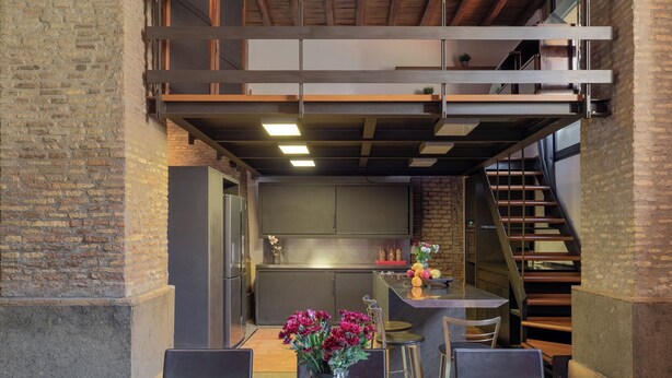 A modern kitchen with a central island, a dining area with a table and chairs, and a staircase leading to an upper level. - Santa Cecilia Loft (Rome)