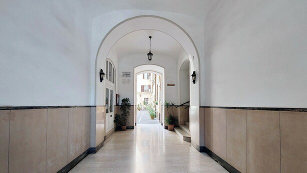 A corridor with white walls, beige paneling, and a marble floor. There are potted plants along the sides and a sign on the wall. - Cozy Merulana (Rome)