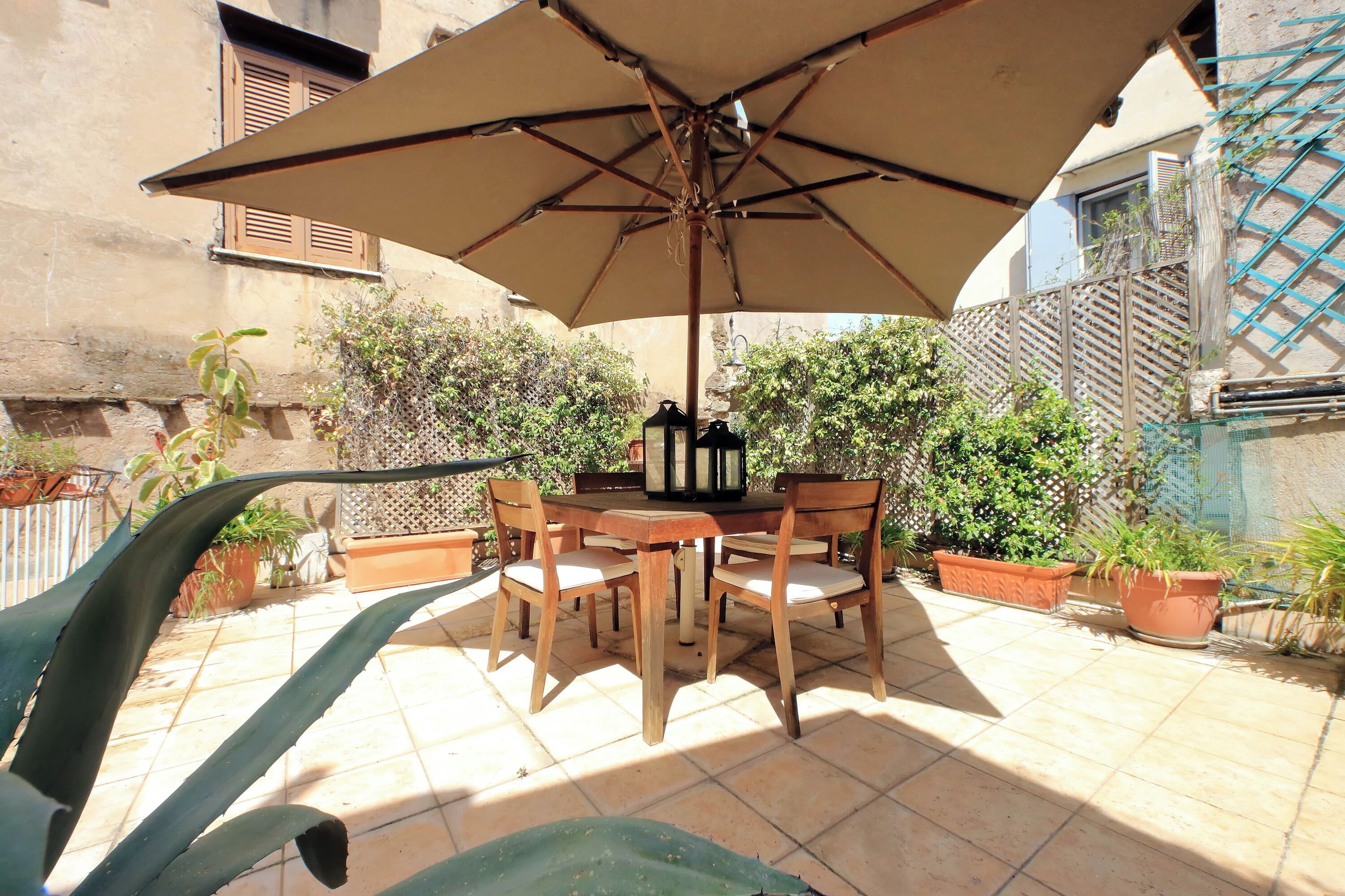 Terrassa exterior amb taula i cadires sota un parasol gran, envoltada de plantes en testos i un paviment de rajoles.