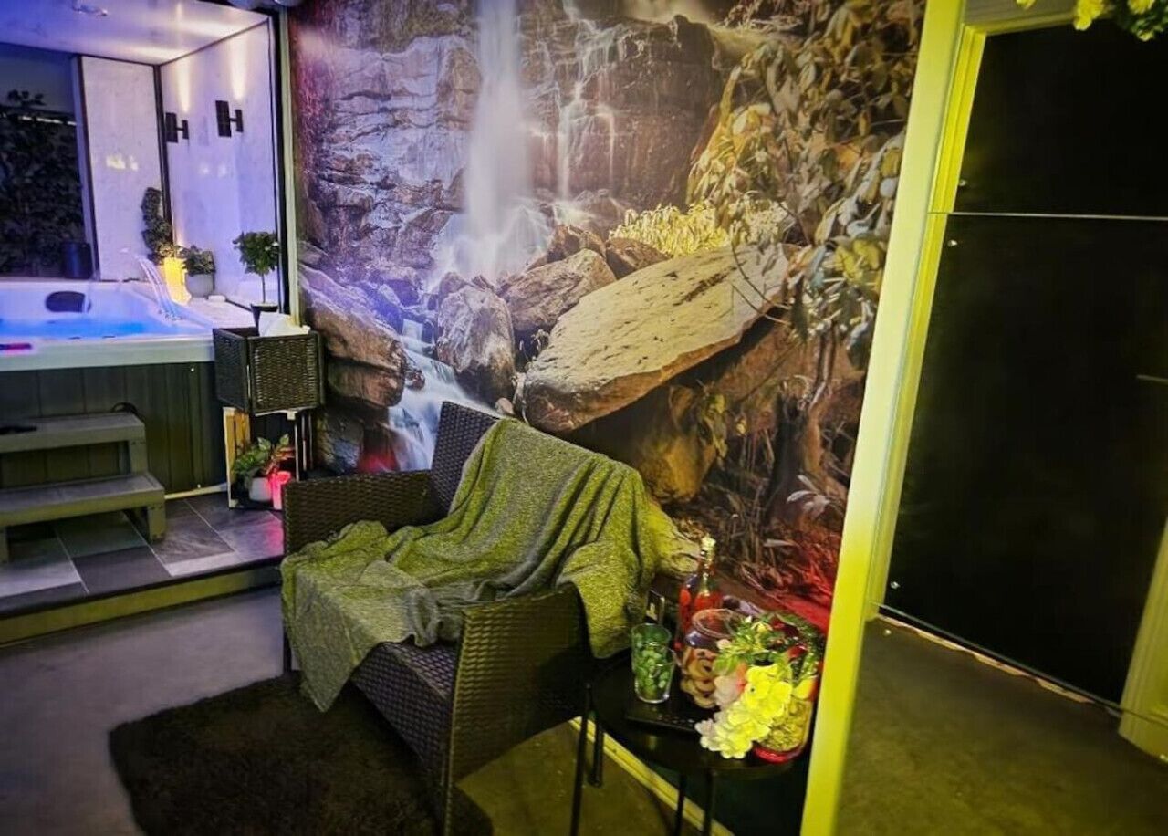 Ein gemütliches Zimmer mit einer großen Wandmalerei eines Wasserfalls, einem bequemen Sessel, einem kleinen Tisch mit Getränken und einer Whirlwanne im Hintergrund.